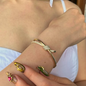 May include: A gold-coloured bracelet featuring a knot design, embellished with small, sparkling details. The bracelet is worn on a wrist, highlighting its elegant style. The nails are painted with nail art.