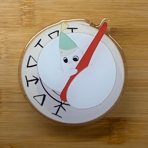 May include: A circular acrylic charm featuring a white ghost character wearing a small green party hat. The ghost is holding a large red pen against a white background with a circular border containing black geometric symbols and markings.
