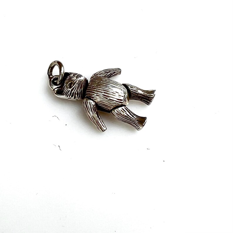 May include: A silver-colored teddy bear charm with articulated limbs. The bear is lying down on a white surface. The charm has a loop at the top for attaching to a necklace or bracelet.