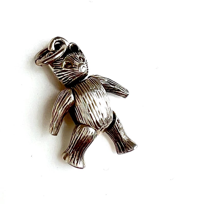 May include: A silver-colored teddy bear charm with articulated limbs. The bear has etched details and a small loop at the top for attachment to a necklace or bracelet. The charm is set against a white background.