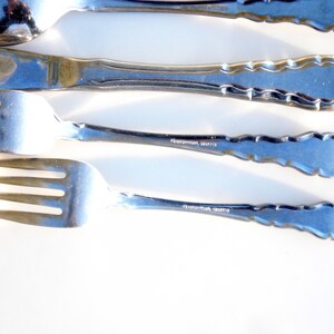 Puede incluir: Cuatro piezas de cubiertos, incluyendo un tenedor, un cuchillo, una cuchara y un tenedor m&aacute;s peque&ntilde;o, est&aacute;n dispuestos en una fila sobre una superficie blanca. Los cubiertos est&aacute;n hechos de acero inoxidable pulido y tienen un borde decorativo festoneado. La parte posterior de cada pieza de cubiertos est&aacute; estampada con las palabras "International, Stainless".