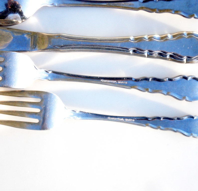 Puede incluir: Cuatro piezas de cubiertos, incluyendo un tenedor, un cuchillo, una cuchara y un tenedor m&aacute;s peque&ntilde;o, est&aacute;n dispuestos en una fila sobre una superficie blanca. Los cubiertos est&aacute;n hechos de acero inoxidable pulido y tienen un borde decorativo festoneado. La parte posterior de cada pieza de cubiertos est&aacute; estampada con las palabras "International, Stainless".