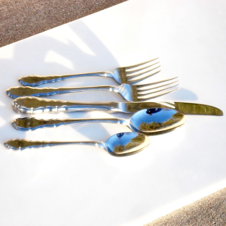 Puede incluir: Un juego de cinco cubiertos plateados, que incluye un tenedor, un cuchillo y tres cucharas, dispuestos sobre una superficie blanca.