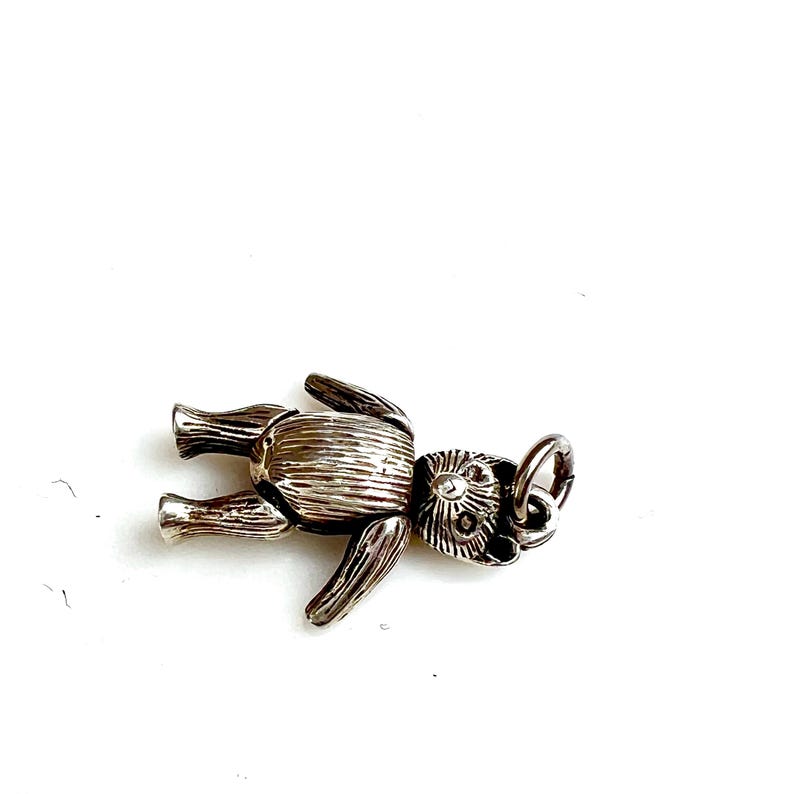 May include: A silver-colored teddy bear charm with detailed features, including articulated limbs and a textured body. The charm has a small loop for attaching to a bracelet or necklace. The bear is lying on its back.