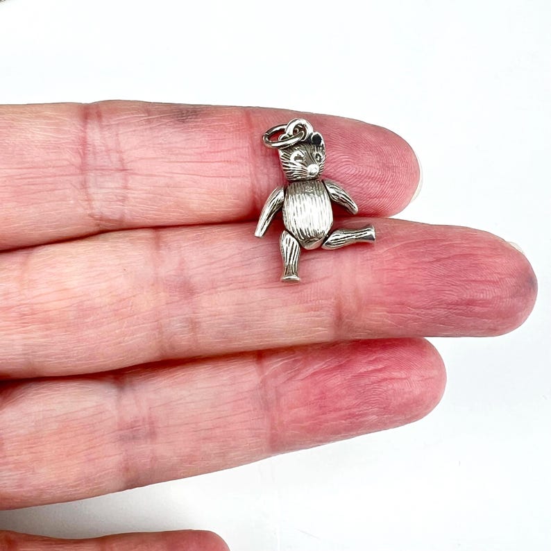 May include: A small, silver-colored teddy bear charm with articulated limbs. The bear is posed with one leg raised, and has a loop at the top for attaching to a bracelet or necklace. The charm is detailed with etched fur and facial features.