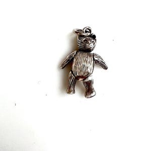 May include: A silver-colored teddy bear charm with articulated limbs and a textured surface. The bear has a loop at the top for attaching to a bracelet or necklace. The charm is set against a white background.