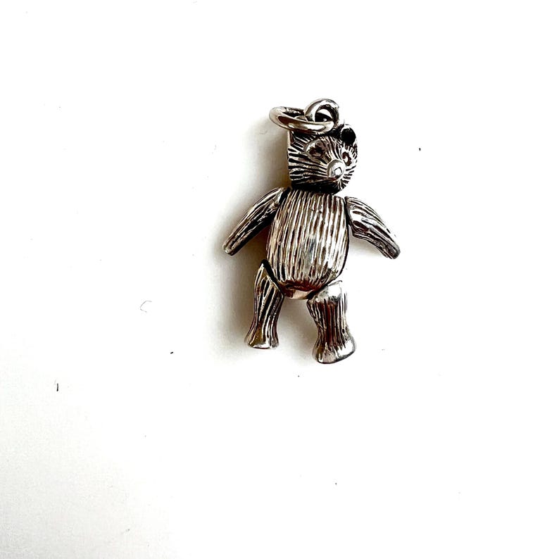 May include: A silver-colored teddy bear charm with articulated limbs and a textured surface. The bear has a loop at the top for attaching to a bracelet or necklace. The charm is set against a white background.