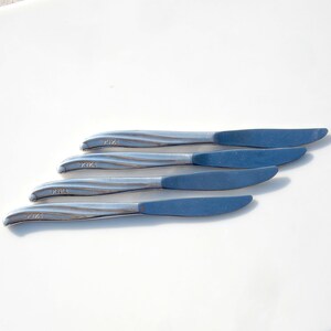 May include: Four silver butter knives with blue handles. The knives are arranged in a row, with the handles facing up. The knives are all marked with the letters "TWA" and a number.