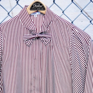 May include: A white and burgundy striped blouse with a bow tie. The blouse has a collar and long sleeves. The tag on the blouse reads "Strambergs" and "Paris".
