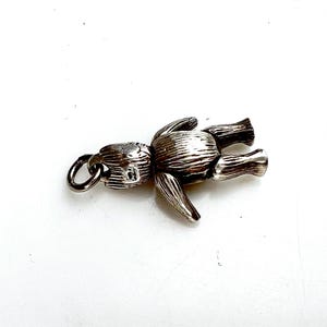 May include: A silver-colored articulated teddy bear charm with a ring for attachment. The bear has detailed features, including a head, torso, arms, and legs. The charm is lying on a white surface.