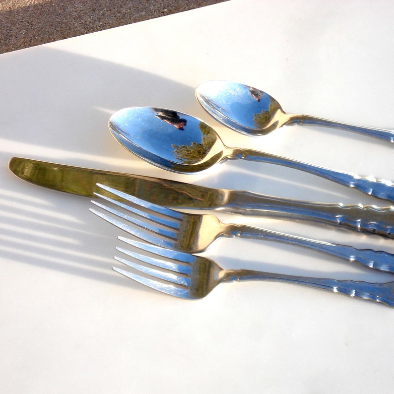 Puede incluir: Un juego de cubiertos, que incluye un cuchillo, un tenedor y dos cucharas, dispuestos sobre una superficie blanca. Los cubiertos est&aacute;n pulidos y reflejan la luz.