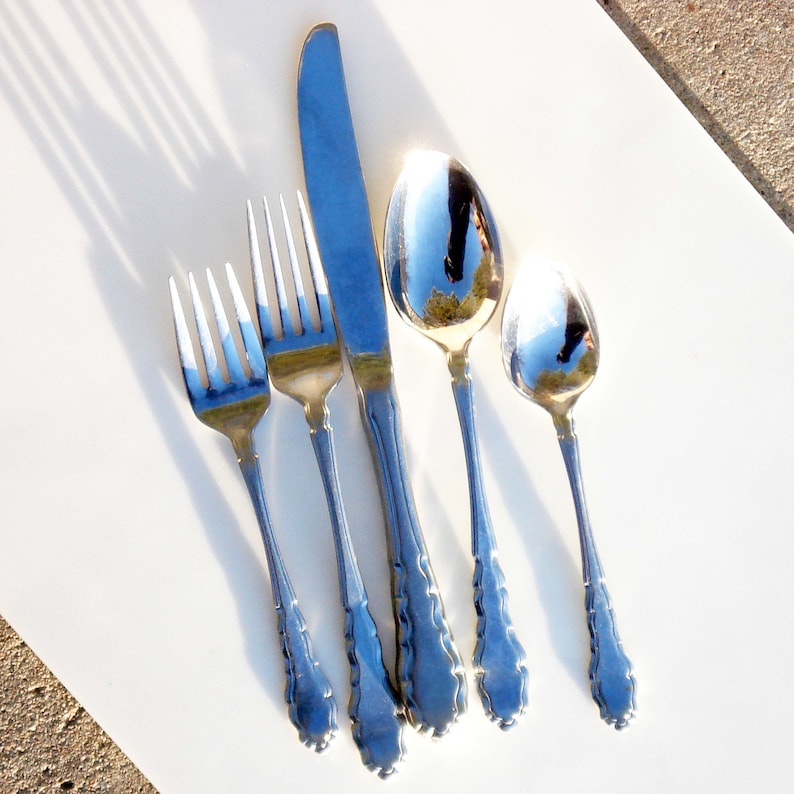 Puede incluir: Un juego de cinco cubiertos plateados, que incluye un cuchillo, dos tenedores y dos cucharas. Los cubiertos est&aacute;n dispuestos sobre una superficie blanca.