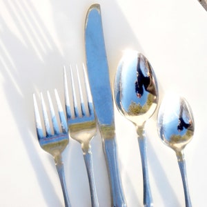 Puede incluir: Un juego de cuatro cubiertos plateados, que incluye un cuchillo, dos tenedores y una cuchara. Los cubiertos est&aacute;n dispuestos verticalmente sobre una superficie blanca, con el cuchillo en el centro y los tenedores a cada lado. La cuchara est&aacute; a la derecha del cuchillo. Los cubiertos reflejan la luz, creando un efecto brillante.