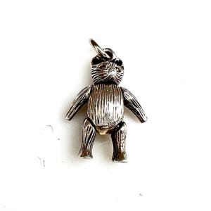 May include: A silver teddy bear charm with a textured, ribbed body and articulated limbs. The bear has a small loop at the top for attaching to a necklace or bracelet. The charm is set against a white background.
