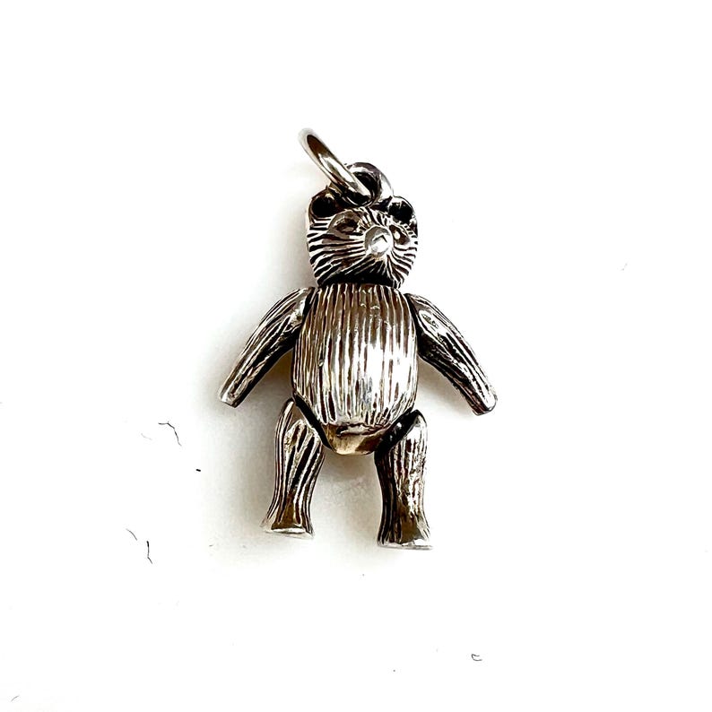 May include: A silver teddy bear charm with a textured, ribbed body and articulated limbs. The bear has a small loop at the top for attaching to a necklace or bracelet. The charm is set against a white background.