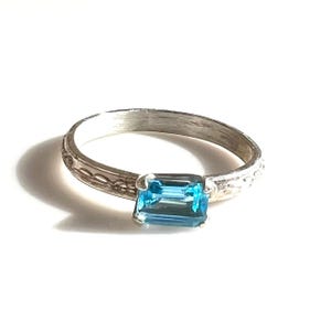 May include: A silver ring with an emerald-cut, bright blue gemstone. The band has a textured, etched design. The ring is set against a white background, highlighting the gemstone's color and the ring's details.
