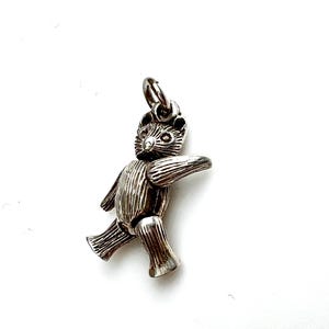 May include: A silver-toned charm in the shape of a bear, with articulated limbs and a textured surface. The bear is depicted in a walking pose, with one arm extended. The charm has a loop at the top for attachment to a bracelet or necklace.