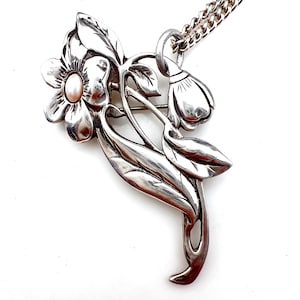 May include: A silver-toned floral brooch and pendant necklace. The brooch features a large flower with a pearl center, leaves, and a bud. The necklace has a delicate chain. The jewelry is likely vintage or antique.