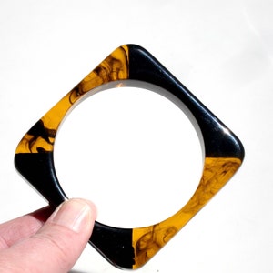 May include: Square bangle bracelet with a geometric design. The bracelet is divided into two colors: black and a marbled amber. The bracelet is held by a hand against a white background. The bracelet has a square shape with rounded corners.