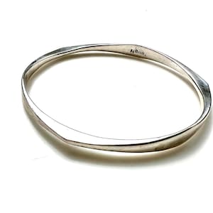 May include: A silver-colored bangle bracelet with a smooth, polished finish and a slightly irregular, angular shape. The inner surface of the metal band features a clear, stamped inscription that reads 800.