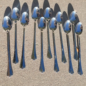 May include: A collection of ten shiny, silver-colored spoons with long, slender handles. The spoons have a teardrop-shaped bowl and a decorative, fan-like design at the base of the handle. The spoons are arranged on a textured, light-colored surface.
