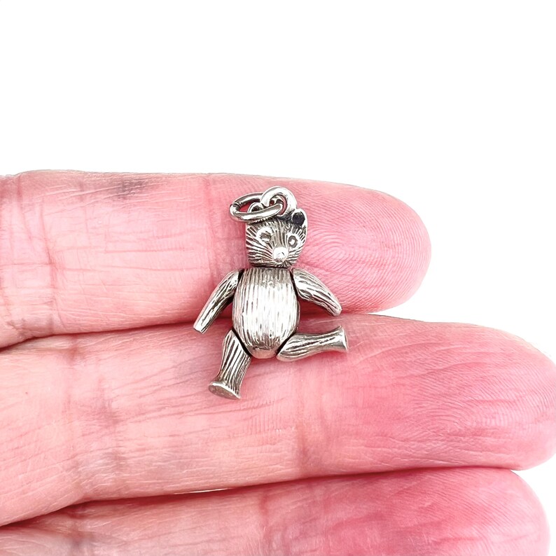 May include: A small, silver-colored teddy bear charm with articulated limbs. The bear is sitting with one leg extended and has a loop at the top for attaching to a bracelet or necklace. The bear's features include small ears and a textured body.