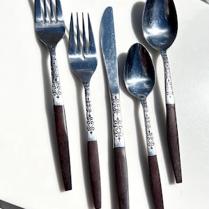 May include: A set of vintage silverware including forks, spoons, and a knife. The stainless steel utensils have dark brown handles and decorative silver accents. The spoons and forks are of different sizes.