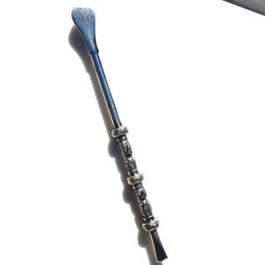 May include: A silver-colored yerba mate bombilla with a blue-toned head featuring small holes. The stem is long and slender, with decorative bands near the bottom. The bombilla is designed for filtering yerba mate.