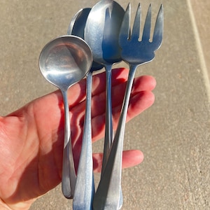 May include: A set of four stainless steel utensils: a ladle, two spoons, and a fork. The silverware has a simple, classic design with long, slender handles and rounded or curved heads. The utensils are held in a hand.