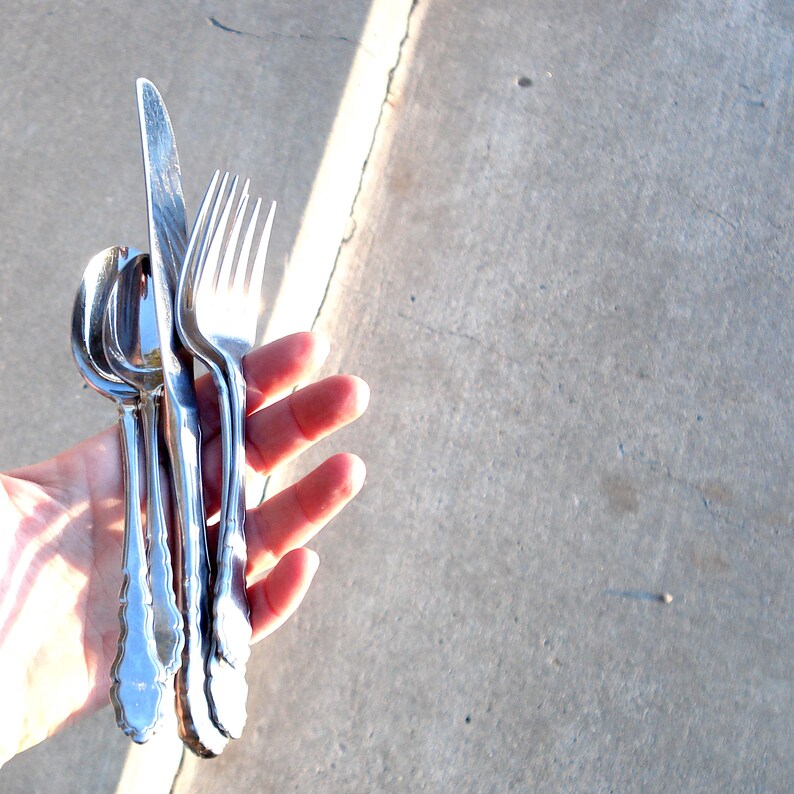 Puede incluir: Un juego de cuatro cubiertos de plata, que incluye un cuchillo, un tenedor y dos cucharas, sostenidos en una mano sobre un fondo de hormig&oacute;n gris.