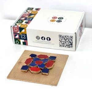 Può includere: Una scatola di cartone bianca con un motivo geometrico colorato, icone dei social media e un codice QR, posizionata dietro una tavola di legno quadrata con un mosaico di piastrelle geometriche rosse e blu a forma di stella.