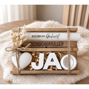 Könnte beinhalten: Eine hölzerne Hochzeitsdekoration mit dem Wort "JA" in weißen Blockbuchstaben. Die Dekoration enthält ein weißes Herz, Trockenblumen und den Text "ALLES LIEBE ZUR Hochzeit" und "WUNSCHERFÜLLER". Die Artikel sind mit Bindfaden zusammengebunden.