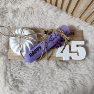 May include: A wooden sign with the text "Happy Birthday" and the number "45" in white. A purple candle with the text "Happy Birthday" is tied to the sign with twine and lavender flowers. A white ceramic candle holder is also on the sign.