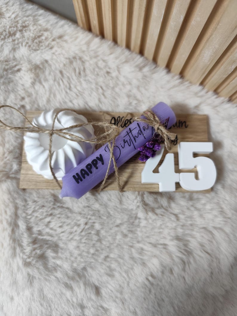 May include: A wooden sign with the text "Happy Birthday" and the number "45" in white. A purple candle with the text "Happy Birthday" is tied to the sign with twine and lavender flowers. A white ceramic candle holder is also on the sign.