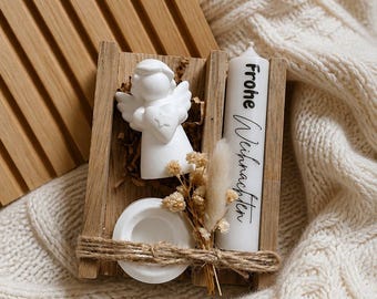 May include: A wooden tray containing a white angel figurine holding a heart, a white candle with the black text Frohe Weihnachten, a small white circular base, and a bundle of dried flowers, all secured together with a piece of brown twine.