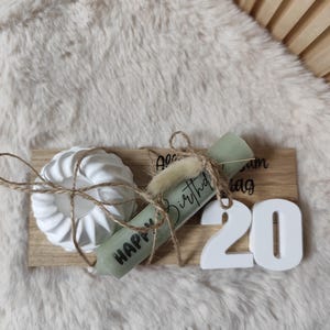 May include: A wooden tray with a white candle holder, a green candle with the text "Happy Birthday", a white scroll with the text "Happy Birthday", and white numbers "20" on a white furry surface.