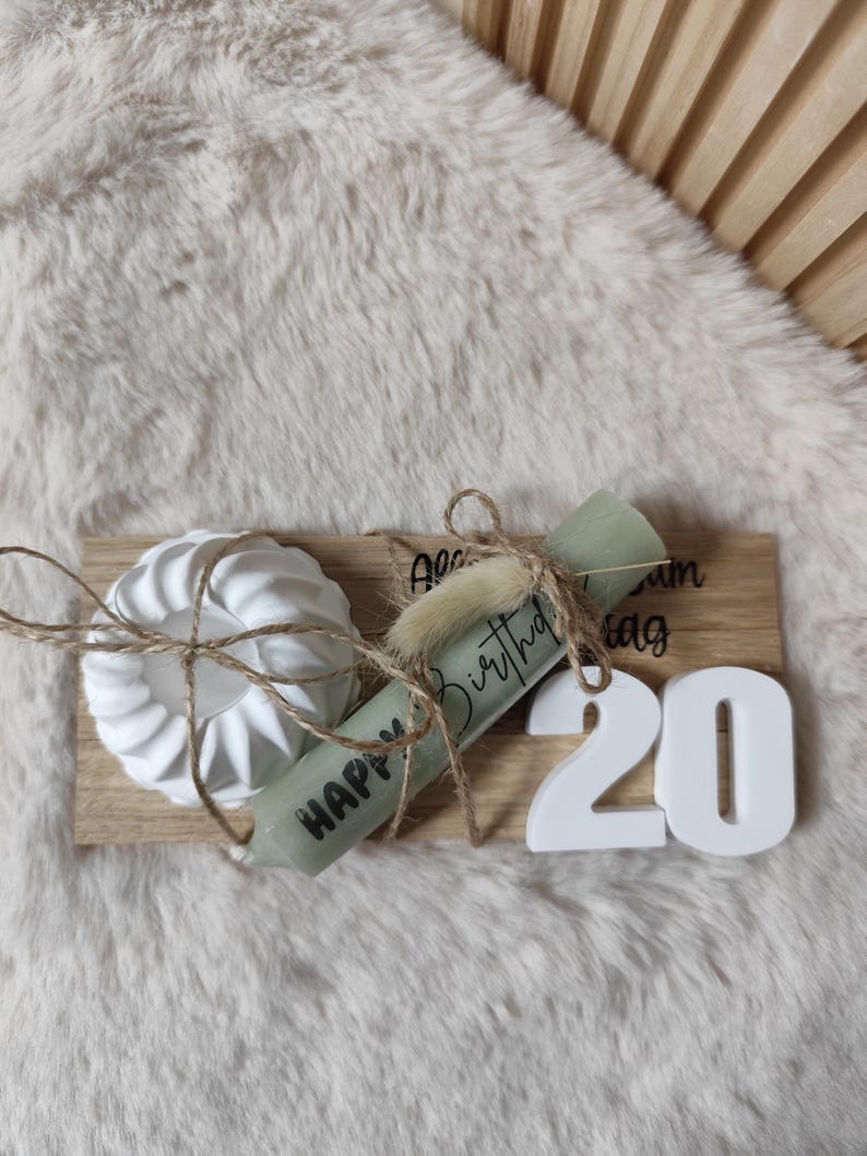 May include: A wooden tray with a white candle holder, a green candle with the text "Happy Birthday", a white scroll with the text "Happy Birthday", and white numbers "20" on a white furry surface.