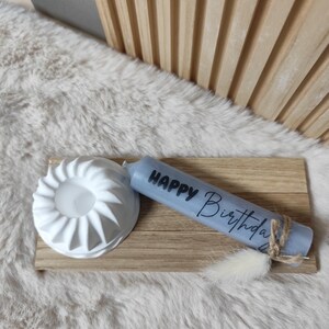 May include: A blue candle with the text "Happy Birthday" on it. The candle is sitting on a wooden tray with a white ceramic candle holder.