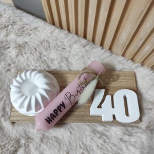 May include: A pink candle with the text "Happy Birthday" on it, a white ceramic candle holder, and white wooden number 40 on a wooden board.