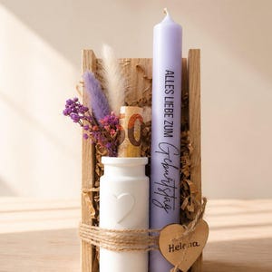 Könnte beinhalten: Eine hellviolette Stabkerze mit dem Text ALLES LIEBE ZUM GEBURTSTAG, eine weiße Keramikvase mit Herz, getrocknete violette und cremefarbene Blumen sowie ein gerollter Geldschein in einer Holzbox mit einem Holzherzanhänger namens Helena.