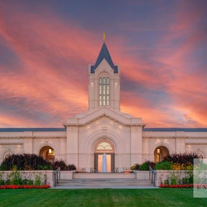 Può includere: Un grande edificio bianco con un'alta torre centrale e un ingresso ad arco, sullo sfondo di un vibrante cielo al tramonto con rosa e arancione. Erba verde e paesaggistica incorniciano la facciata dell'edificio.
