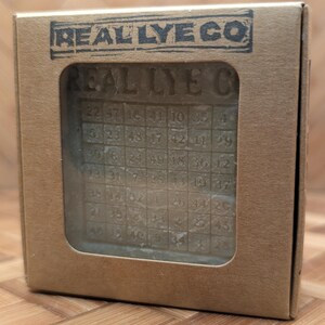May include: A bar of soap with a grid pattern and numbers, visible through a rectangular window in a cardboard box. The box is stamped with "REAL LYE CO" in a dark blue ink. The soap has the words "REAL LYE C" carved into it.