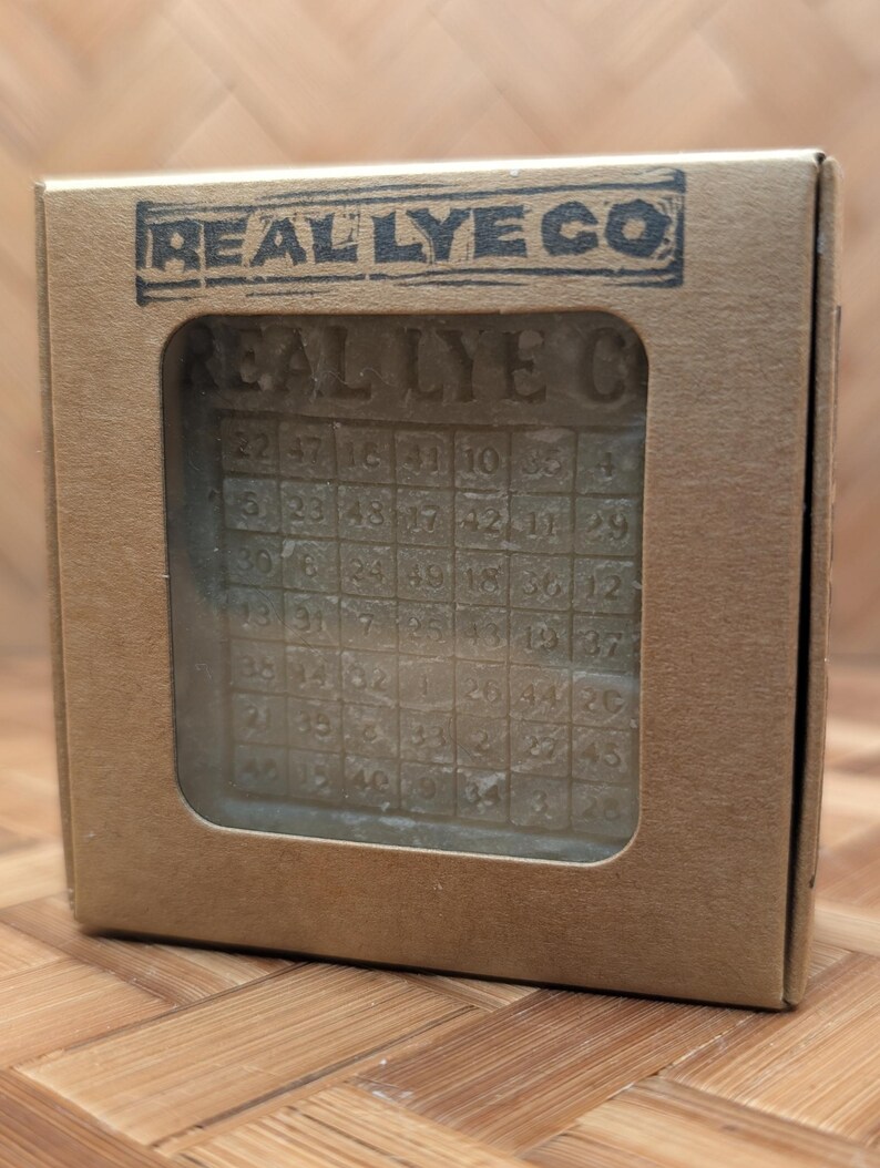 May include: A bar of soap with a grid pattern and numbers, visible through a rectangular window in a cardboard box. The box is stamped with "REAL LYE CO" in a dark blue ink. The soap has the words "REAL LYE C" carved into it.