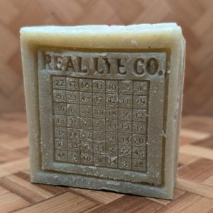 May include: A rectangular bar of soap with the text "REAL LYE CO." and a grid of numbers. The soap is a light green color and sits on a wooden surface. The numbers are arranged in a grid pattern.