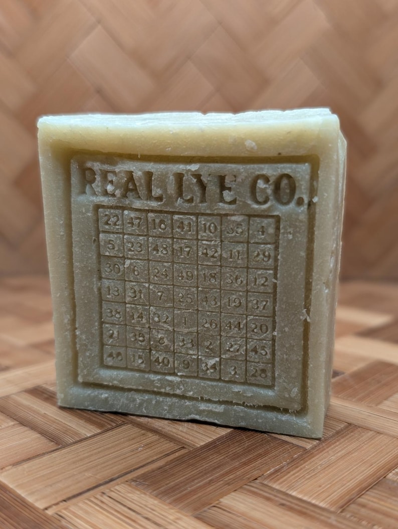 May include: A rectangular bar of soap with the text "REAL LYE CO." and a grid of numbers. The soap is a light green color and sits on a wooden surface. The numbers are arranged in a grid pattern.
