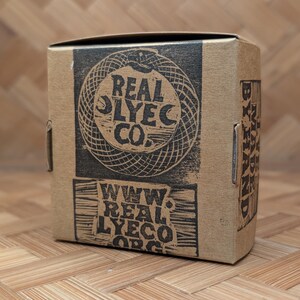 May include: A brown cardboard box with black printed text and graphics. The box features the words "REAL LYECO CO." and "WWW. REAL LYECO.ORG". The box is sitting on a bamboo surface.