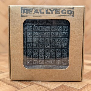 May include: A dark gray soap bar with a grid of numbers, displayed in a cardboard box. The box has the text "REALLYE CO" printed on it. The soap has a matrix of numbers from 1 to 49.