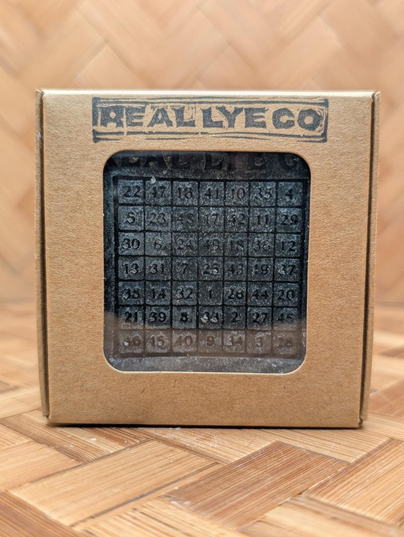May include: A dark gray soap bar with a grid of numbers, displayed in a cardboard box. The box has the text "REALLYE CO" printed on it. The soap has a matrix of numbers from 1 to 49.
