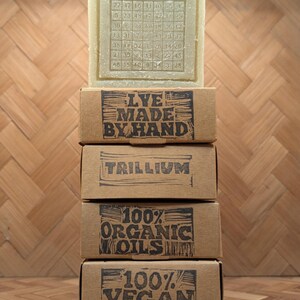 May include: A stack of four brown cardboard boxes with black text, topped by a bar of soap. The soap is stamped with "REAL LYE CO." and a grid of numbers. The boxes read "LYE MADE BY HAND", "TRILLIUM", "100% ORGANIC OILS", and "100% VEGAN".