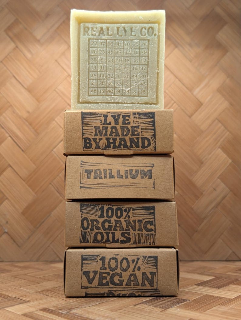 May include: A stack of four brown cardboard boxes with black text, topped by a bar of soap. The soap is stamped with "REAL LYE CO." and a grid of numbers. The boxes read "LYE MADE BY HAND", "TRILLIUM", "100% ORGANIC OILS", and "100% VEGAN".